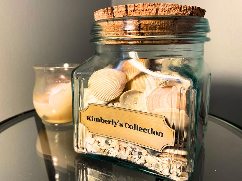 May include: A square, light blue-tinted glass jar with a cork lid, filled with various white and tan seashells and small fragments. A rectangular gold-toned label on the front features the black printed text, Kimberly's Collection.