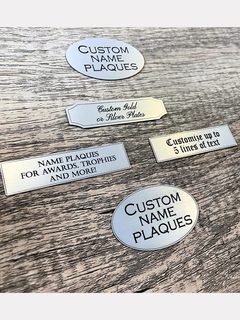 May include: A collection of silver-colored metal name plaques in oval and rectangular shapes. The plaques feature black engraved text including CUSTOM NAME PLAQUES, Custom Gold or Silver Plates, Customize up to 5 lines of text, and NAME PLAQUES FOR AWARDS, TROPHIES AND MORE!