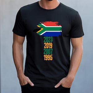 Może przedstawiać: Czarny T-shirt z nadrukiem przetartej flagi Republiki Południowej Afryki i lat 2023, 2019, 2007 i 1995 pod spodem.