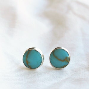 May include: A pair of round stud earrings with turquoise-colored centers and silver-colored metal settings. The turquoise has a gold-colored vein pattern. The earrings are displayed on a white, textured surface.