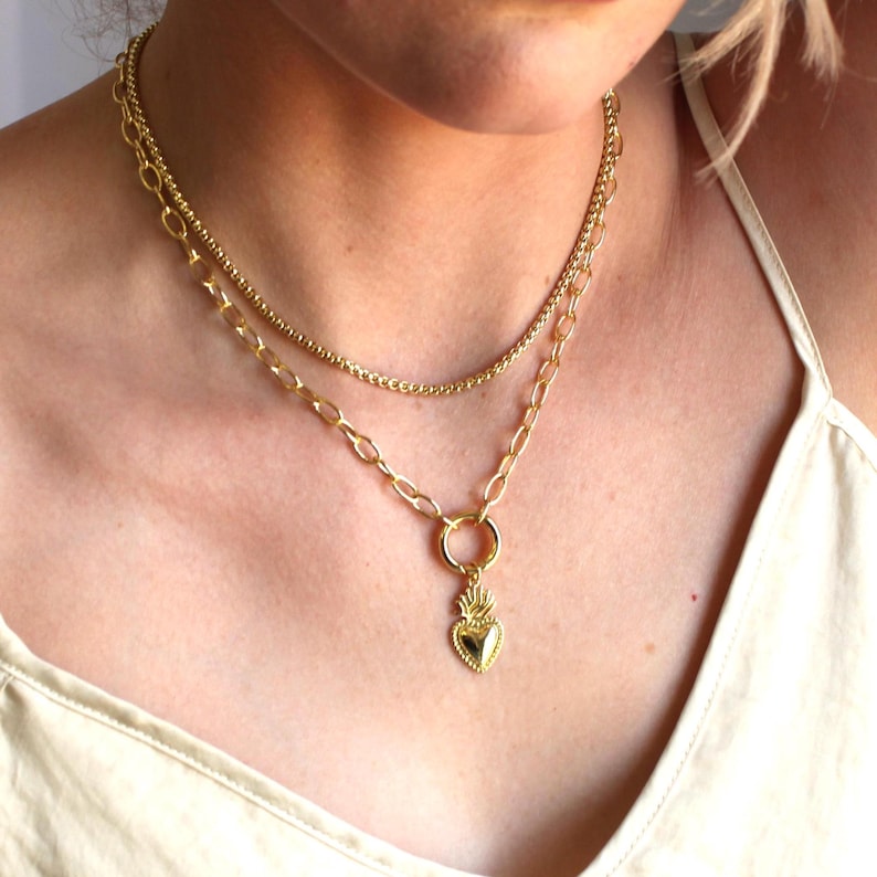 May include: Gold layered necklace with a heart pendant. The heart pendant has a detailed design with a flame-like pattern.