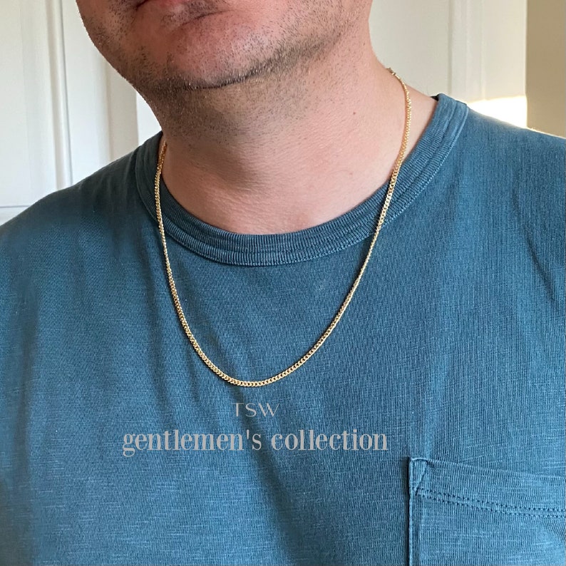 Pu&ograve; includere: Una collana a catena d'oro su una persona che indossa una camicia blu turchese. La catena &egrave; di stile curb e il testo "TSW gentlemen's collection" &egrave; visibile sotto la catena.