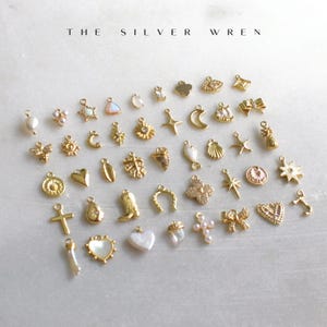 K&ouml;nnte beinhalten: Eine Sammlung goldfarbener Charms mit verschiedenen Designs, darunter Herzen, Sterne, Monde, Kreuze und Blumen. Die Charms sind auf einer wei&szlig;en Oberfl&auml;che angeordnet, oben steht der Text "THE SILVER WREN".