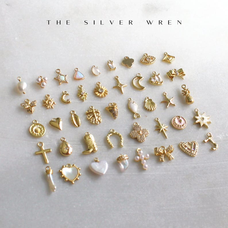 K&ouml;nnte beinhalten: Eine Sammlung goldfarbener Charms mit verschiedenen Designs, darunter Herzen, Sterne, Monde, Kreuze und Blumen. Die Charms sind auf einer wei&szlig;en Oberfl&auml;che angeordnet, oben steht der Text "THE SILVER WREN".