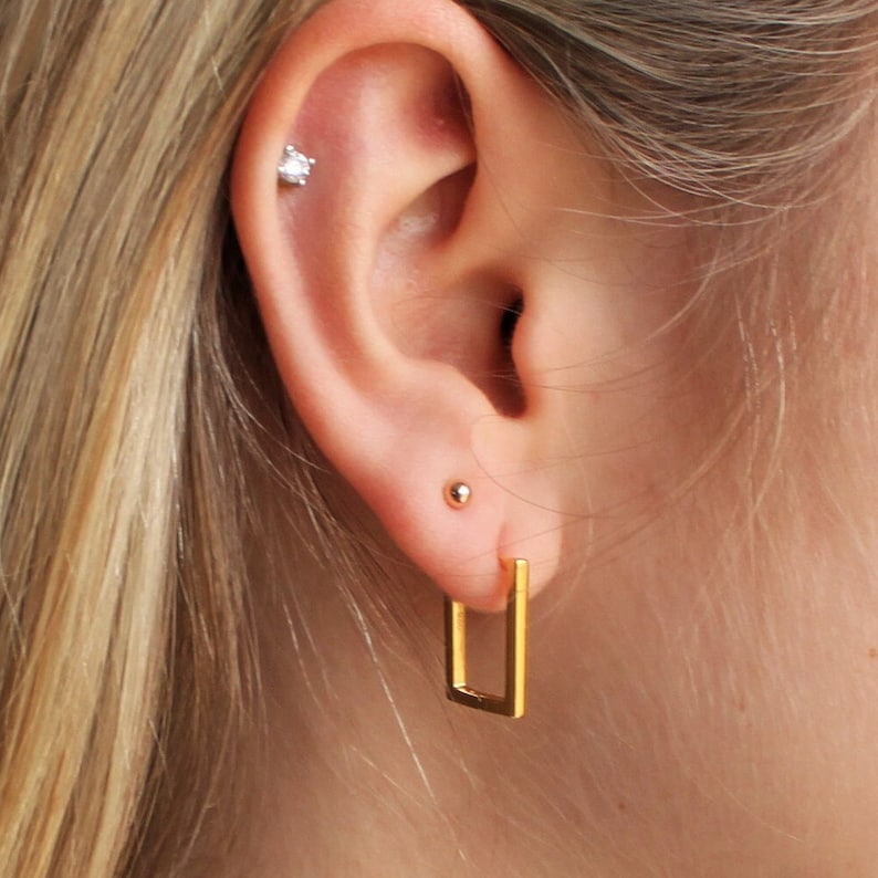 May include: Close-up of an ear with three earrings. A small, clear, round stud is in the upper ear, a small gold ball stud is in the middle, and a gold rectangular hoop earring is in the lower lobe.
