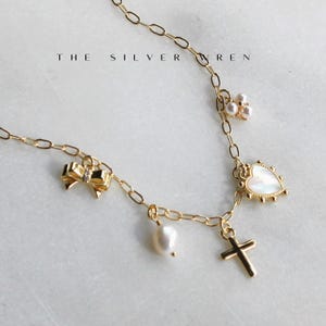 K&ouml;nnte beinhalten: Eine goldene Kettenkette mit mehreren Anh&auml;ngern, darunter eine Perle, ein Herz, eine Schleife und ein Kreuz. Die Anh&auml;nger sind alle aus Gold und haben ein zartes Design. Die Kette ist eine einfache, zarte Kette.