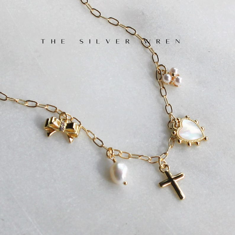K&ouml;nnte beinhalten: Eine goldene Kettenkette mit mehreren Anh&auml;ngern, darunter eine Perle, ein Herz, eine Schleife und ein Kreuz. Die Anh&auml;nger sind alle aus Gold und haben ein zartes Design. Die Kette ist eine einfache, zarte Kette.