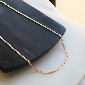 Pu&ograve; includere: Una collana a catena dorata con uno stile a maglie a curb. La catena &egrave; appoggiata su una superficie nera.
