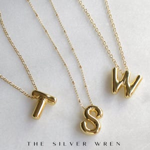 May include: Three gold necklaces with initial pendants. The necklaces feature a T, an S, and a W pendant. The pendants are made of a thick, chunky gold metal.