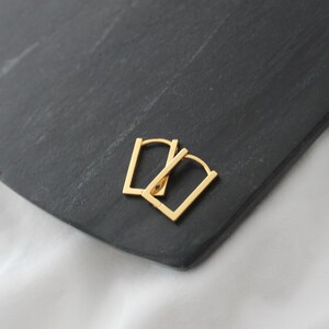 May include: A pair of gold-colored, rectangular hoop earrings. The earrings are displayed on a dark gray, textured surface, with a white background visible in the lower right corner. The earrings have a simple, modern design.