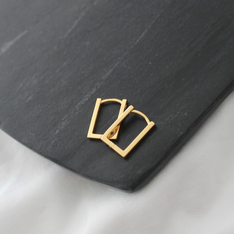 May include: A pair of gold-colored, rectangular hoop earrings. The earrings are displayed on a dark gray, textured surface, with a white background visible in the lower right corner. The earrings have a simple, modern design.