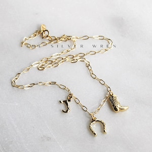 K&ouml;nnte beinhalten: Eine goldene Kettenkette mit drei Anh&auml;ngern: einem Hufeisen, einem Cowboystiefel und dem Buchstaben "J".