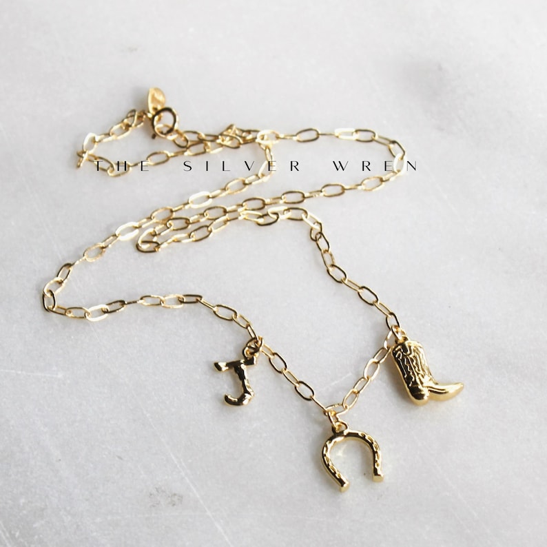 K&ouml;nnte beinhalten: Eine goldene Kettenkette mit drei Anh&auml;ngern: einem Hufeisen, einem Cowboystiefel und dem Buchstaben "J".