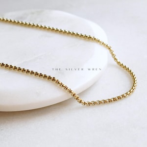 May include: A gold chain necklace with a delicate box chain design. The chain is laying on a white surface.