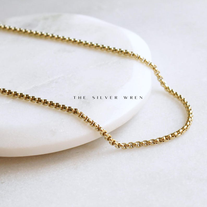 May include: A gold chain necklace with a delicate box chain design. The chain is laying on a white surface.