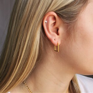 May include: A close-up of a person's ear with a gold rectangular hoop earring. The ear also has a small stud earring and a small, sparkly stud in the upper cartilage. The person has long, blonde hair and a gold chain necklace.