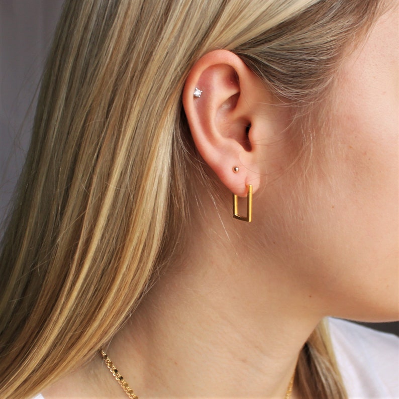 May include: A close-up of a person's ear with a gold rectangular hoop earring. The ear also has a small stud earring and a small, sparkly stud in the upper cartilage. The person has long, blonde hair and a gold chain necklace.