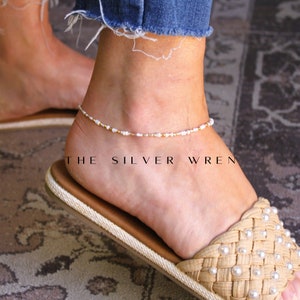 May include: Ankle bracelet with white and gold beads. The delicate jewelry is worn on an ankle, with a woven sandal in the foreground. The text "THE SILVER WREN" is printed on the image.