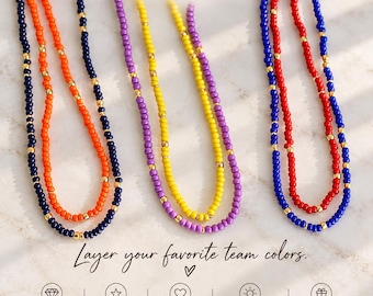 May include: Three beaded necklaces featuring small round beads in dark blue, orange, purple, yellow, and red, interspersed with tiny gold-tone metallic spacers. The text THE SILVER WREN and Layer your favorite team colors. appear on the image.