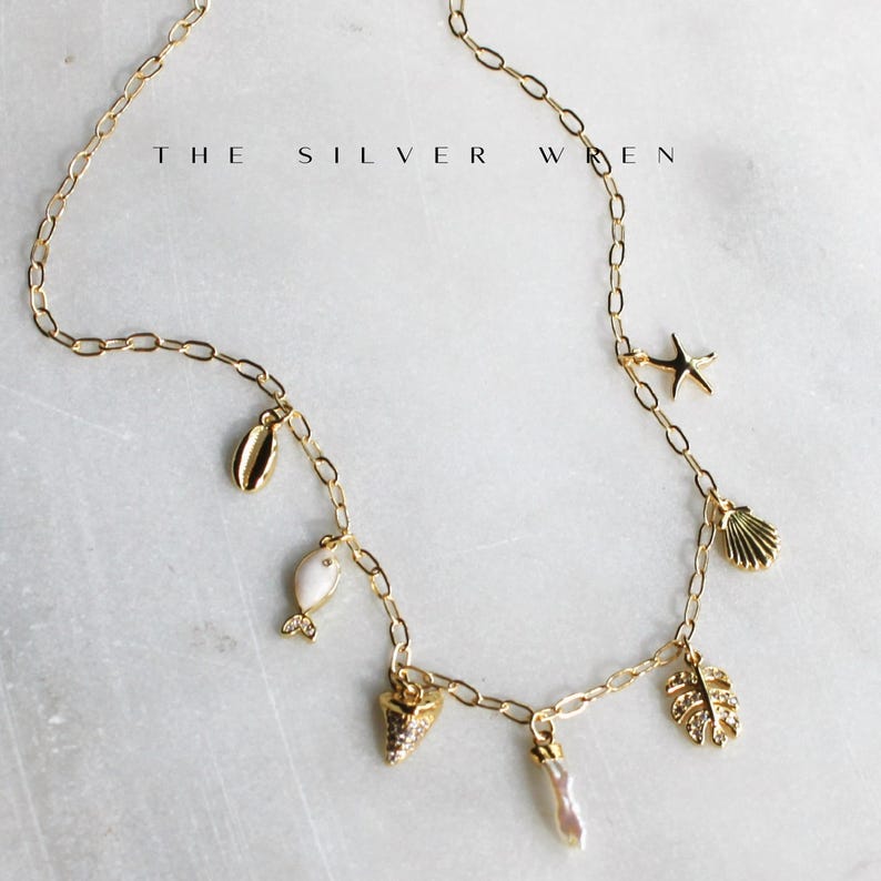 K&ouml;nnte beinhalten: Goldene Kettenkette mit mehreren Anh&auml;ngern, darunter ein Seestern, eine Muschel, ein Fisch und ein Blatt. Die Anh&auml;nger sind aus wei&szlig;em und goldenem Metall. Der Text "THE SILVER WREN" ist im Hintergrund sichtbar.