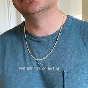 Pu&ograve; includere: Una collana a catena d'oro su una persona che indossa una camicia blu turchese. La catena &egrave; di stile curb e il testo "TSW gentlemen's collection" &egrave; visibile sotto la catena.