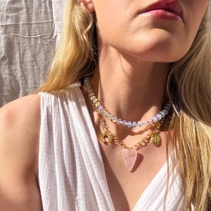 May include: A layered necklace with iridescent beads, a gold chain with a heart-shaped pink stone pendant, and a gold charm.
