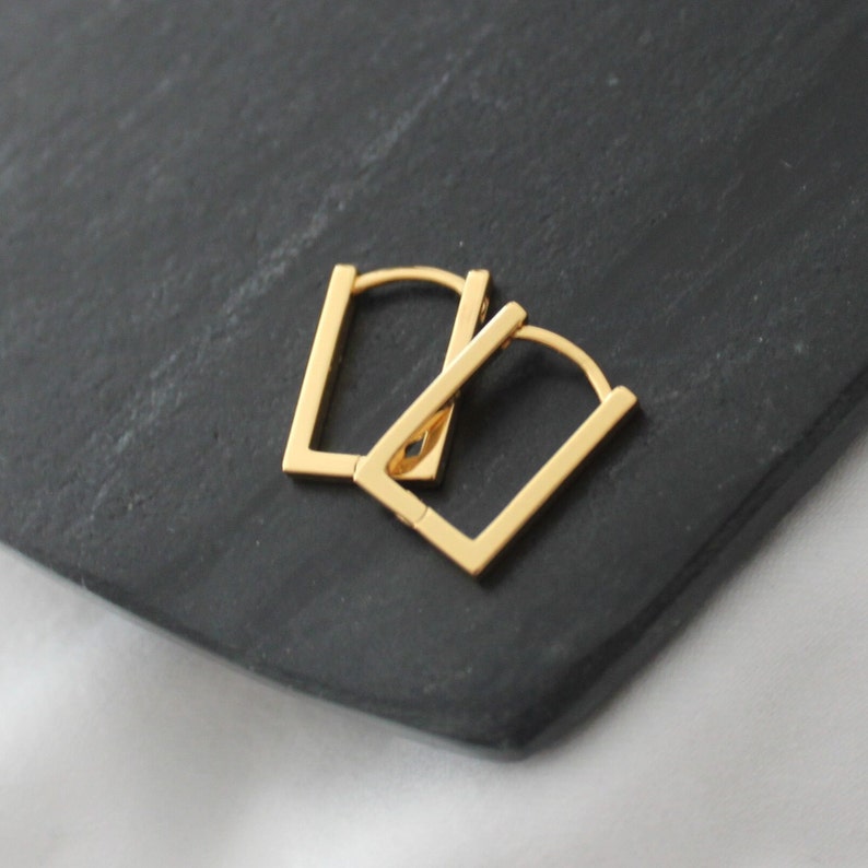 May include: A pair of gold-colored square hoop earrings. The earrings are displayed on a dark gray surface. The earrings have a simple, geometric design.