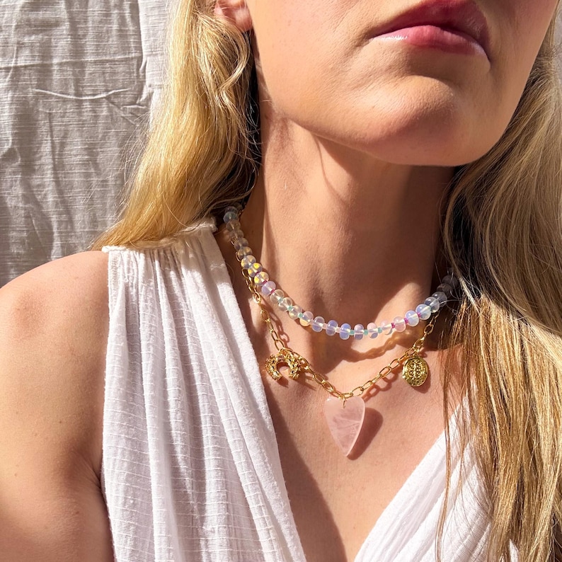 May include: A layered necklace with iridescent beads, a gold chain with a heart-shaped pink stone pendant, and a gold charm.