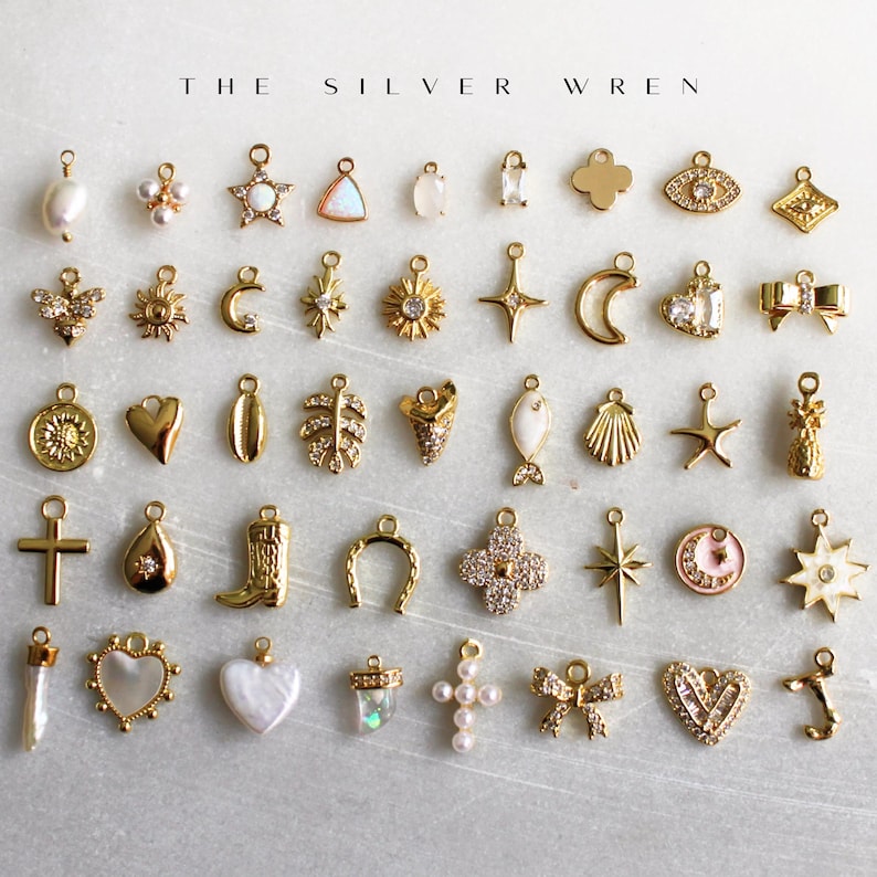 K&ouml;nnte beinhalten: Eine Sammlung von 36 goldfarbenen Charms mit verschiedenen Designs, darunter Herzen, Sterne, Kreuze, Monde und vieles mehr. Die Charms sind in Reihen auf einer wei&szlig;en Oberfl&auml;che angeordnet. Der Text "THE SILVER WREN" ist oben im Bild sichtbar.
