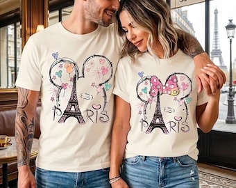 Könnte beinhalten: Zwei cremefarbene T-Shirts mit einer Grafik des Eiffelturms in Paris. Das Design umfasst eine Mickey Mouse-Kopfkontur, Feuerwerk und das Wort "PARIS". Die Shirts werden mit blauen Jeans getragen.