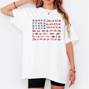 May include: A white short-sleeve t-shirt featuring a graphic design shaped like a flag. The canton consists of blue starfish, while the stripes are composed of various red marine life illustrations, including crabs, lobsters, and fish.