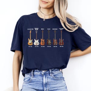 May include: A navy blue t-shirt displaying six guitar illustrations with names and production years: Alligator, Travis Bean, Wolf, Tiger, Rosebud, and Lightning Bolt. The shirt is worn by a person in blue jeans.