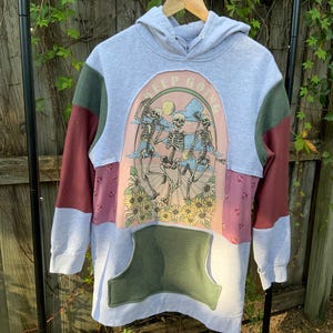 May include: A gray hooded sweatshirt with a patchwork design. The front features a pink panel with a graphic of skeletons dancing with the text "Keep Going". The sleeves are burgundy and green.