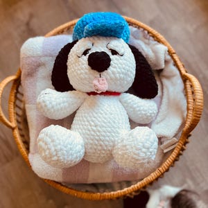 May include: A handmade white crochet dog with black ears and eyes, wearing a blue hat and a red collar. The dog is inside a woven basket on top of a striped blanket. The dog's tongue is sticking out.