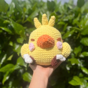 May include: A handmade yellow crochet plush toy shaped like a chick with an orange beak, pink cheeks, and brown and white feet. The plush has three yellow crests on top of its head and closed eyes.