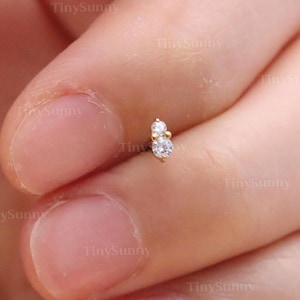 May include: A small, gold-colored earring with two clear, round gemstones. The earring is held between fingers, showcasing its delicate design. The image is well-lit, highlighting the earring's sparkle. The text "@TinySunny" is visible in the image.