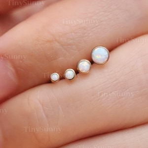 Könnte beinhalten: Ein Satz von fünf goldfarbenen Ohrsteckern, jeweils mit einem runden, weißen Opalstein. Die Ohrringe sind in einer gebogenen Linie angeordnet, wobei die Opalsteine an Größe zunehmen. Die Ohrringe werden auf einem Finger präsentiert.