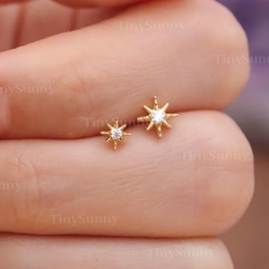 Peut inclure: Une paire de boucles d'oreilles clous en forme d'étoile dorée, l'une légèrement plus grande que l'autre, chacune avec une pierre précieuse claire au centre. Les boucles d'oreilles sont présentées sur une main. Le texte "@TinySunny" est visible.