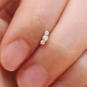 Peut inclure: Un piercing d'oreille incurvé de couleur or, orné de trois pierres précieuses rondes et transparentes. Le bijou est posé sur un fond flou de peau. Le logo TinySunny est visible en bas à droite.