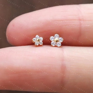Peut inclure: Deux petites boucles d'oreilles à clous en forme de fleur. Chaque boucle d'oreille présente un centre doré entouré de pierres blanches. Les boucles d'oreilles sont présentées sur un doigt, mettant en valeur leur design délicat. Les boucles d'oreilles sont de @Tinysunny Jewelry.