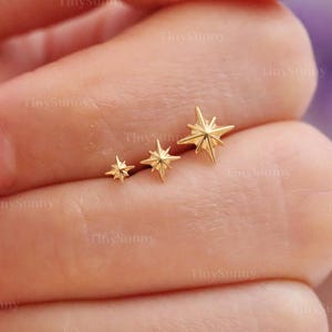 Könnte beinhalten: Drei goldfarbene sternförmige Ohrringe in verschiedenen Größen. Die Ohrringe sind in einer Reihe angeordnet, wobei der kleinste Stern links und der größte rechts ist. Die Ohrringe befinden sich auf den Fingern einer Person. Der Text "@TinySunny" ist sichtbar.