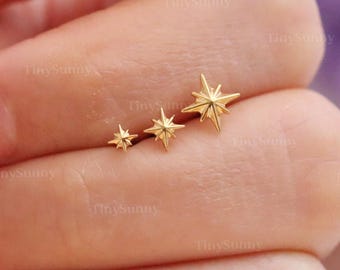 May include: Three gold-colored star-shaped earrings of varying sizes. The earrings are arranged in a row, with the smallest star on the left and the largest on the right. The earrings are on a person's fingers. The text "@TinySunny" is visible.