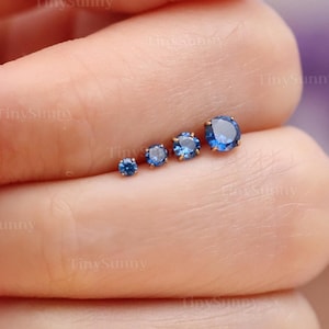 Puede incluir: Cinco pendientes de botón de piedras preciosas azules de diferentes tamaños se muestran en un dedo. Los pendientes son redondos y de color azul brillante. El botón más pequeño está a la izquierda, aumentando de tamaño hacia la derecha.