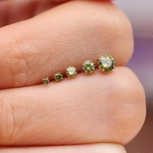 May include: A collection of six small, round, olive-green gemstone studs in varying sizes. The gemstones are set in a light-colored metal, and are displayed on a person's skin. The studs are arranged in a line, showcasing their graduated sizes.