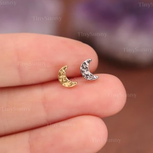 May include: Two small crescent moon earrings, one gold-toned and one silver-toned, are displayed. The earrings have a textured surface and are set against a soft background. The earrings are small and delicate, perfect for everyday wear.