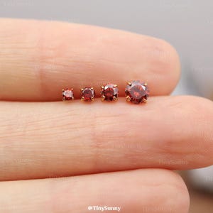 May include: Four round, faceted red gemstone stud earrings arranged in a row by size, from smallest to largest. Each stone is set in a gold-toned metal prong setting, creating a classic look for these small, circular jewelry pieces.