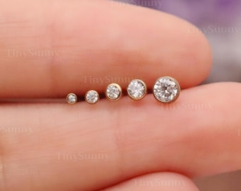 May include: A collection of six gold-colored stud earrings with clear, round gemstones of varying sizes. The earrings are arranged in a row, showcasing their graduated sizes. The image is set against a soft, blurred background.