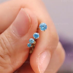 May include: A set of four small, light blue gemstone earrings. The earrings are arranged in a line, with the largest stone at the top. The gemstones are round and set in a gold-colored metal. The earrings are held in a hand.