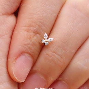 Könnte beinhalten: Ein kleiner, goldfarbener Schmetterlings-Ohrring mit klaren, funkelnden Edelsteinen. Der Ohrring befindet sich auf einem Finger und zeigt sein zartes Design und seine komplizierten Details. Der Schmuck ist ein Ohrstecker.