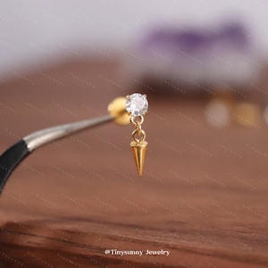May include: A gold-colored earring with a small, clear gemstone and a dangling, pointed charm. The earring is held by a pair of tweezers against a blurred background. The earring is a stud style.